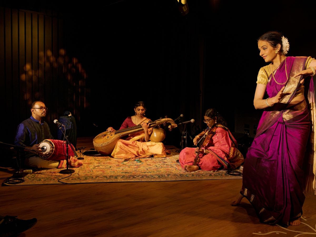 Production photo from And What Will People Say? — full ensemble in Bharatanatyam formation, dramatic stage lighting in deep amber and magenta.